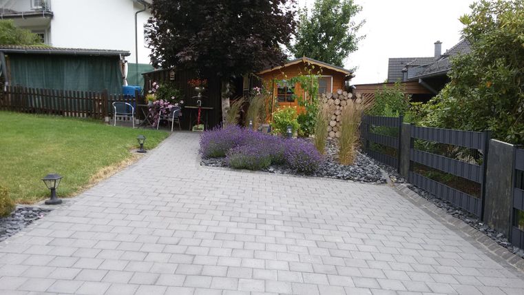 Ein gepflasterter Weg führt durch einen Garten mit Lavendel und verschiedenen Pflanzen. Im Hintergrund sind Holzstapel und Gartenmöbel zu sehen.