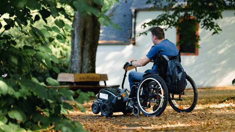 Ein Mann sitzt im Rollstuhl und betrachtet die Umgebung in einem parkähnlichen Bereich. Der Boden ist mit herbstlichen Blättern bedeckt, und im Hintergrund ist ein Gebäude zu sehen.