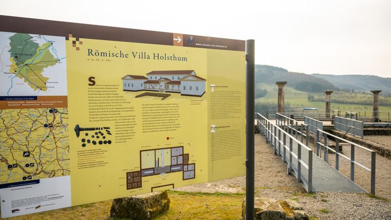 Ein Informationsschild über die Römische Villa Holsthum steht vor den Ruinen des alten Gebäudes. Im Hintergrund sind sanfte Hügel und ein klarer Himmel sichtbar.