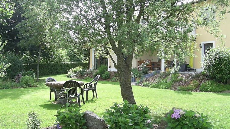 Ein schöner Garten mit einer grünen Wiese und einem großen Baum. Zu sehen sind ein Tisch und Stühle unter dem Baum sowie bunte Blumenbeete.