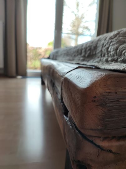 Ein rustikales Holzsofa mit einer natürlichen Oberfläche und klaren Linien. Im Hintergrund sind Fenster mit Tageslicht und eine grüne Aussicht zu sehen.
