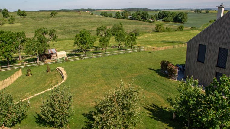 Eine weite grüne Wiese mit Bäumen und einem alten Schuppen. Im Hintergrund sind sanfte Hügel unter blauem Himmel zu sehen.