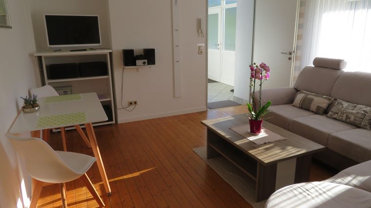 Een lichte woonkamer met een comfortabele bank en een salontafel. Een bureau en een televisie zijn ook zichtbaar, terwijl er verse bloemen op de tafel staan.