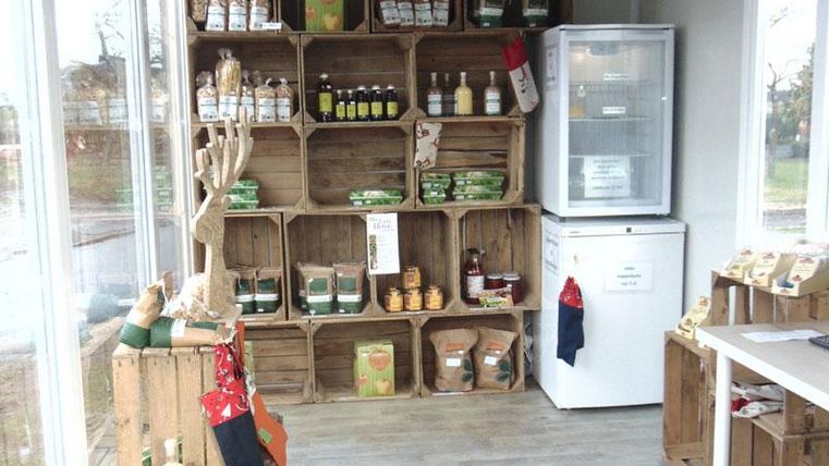Een kleine verkoopsruimte met houten planken, gevuld met verschillende producten. Aan de rechterkant staat een koelkast en een tafel met verdere artikelen.