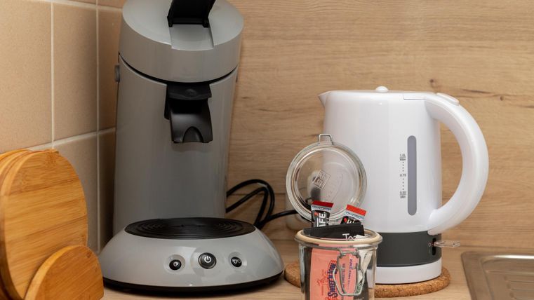 Eine moderne Kaffeemaschine steht neben einem elektrischen Wasserkocher. Auf dem Tisch befindet sich auch ein Glas mit Teebeuteln.
