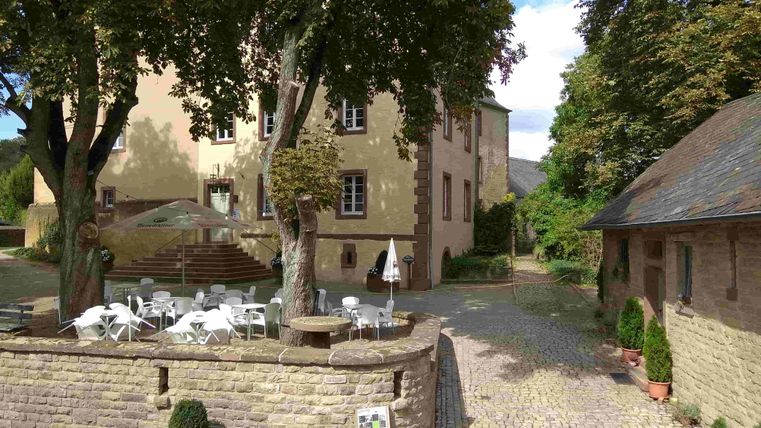 Een rustige binnenplaats met een historisch gebouw en bomen. Tafels en stoelen staan klaar om buiten te ontspannen.