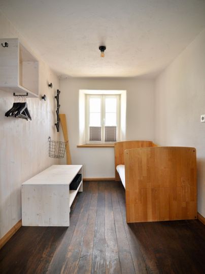 Ein einfaches Zimmer mit einem Holzbett und einem weißen Tisch. Es gibt einen kleinen Schreibtisch und ein Fenster mit Blick nach draußen.