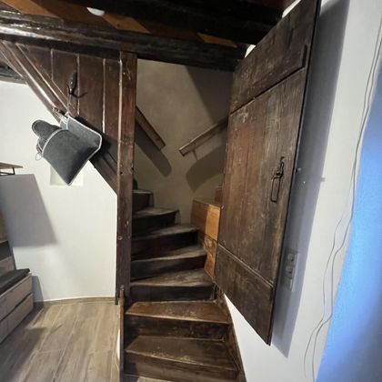 Een smalle houten trap die naar een bovenverdieping leidt. Aan de muur hangt een houten kast met deur.