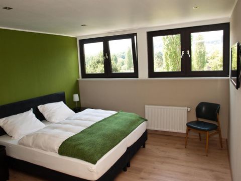 Ein modernes Schlafzimmer mit einem Doppelbett und grüner Bettwäsche. Große Fenster sorgen für viel Tageslicht und einen Blick auf die Natur.