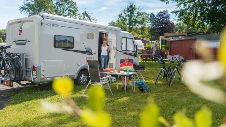 Ein Wohnmobil steht auf einem grünen Campingplatz. Zwei Personen genießen eine Mahlzeit im Freien, umgeben von Bäumen und Fahrrädern.