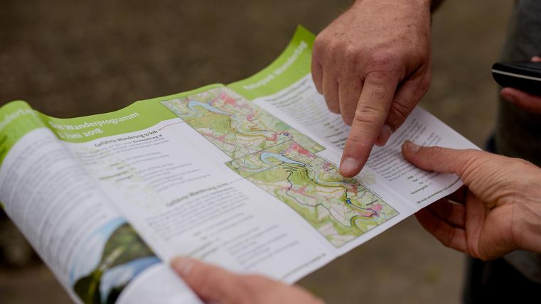 Een persoon wijst met de vinger naar een landkaart. Twee handen houden de kaart vast, terwijl de andere hand een smartphone vasthoudt.