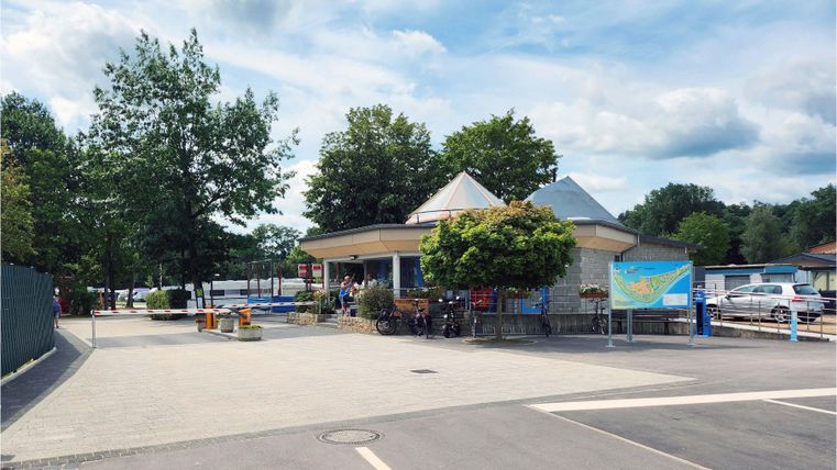 Un bâtiment circulaire avec un grand toit en parasol se trouve dans un environnement calme. Des espaces verts et des arbres entourent la zone, qui invite à se détendre.