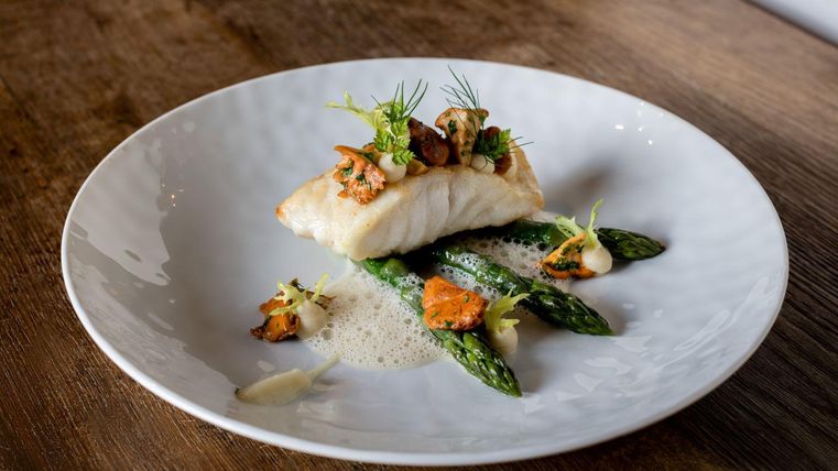 Een mooi opgemaakt bord met gebakken vis, gegarneerd met paddenstoelen en kruiden. Daarbij zijn er verse groene asperges.