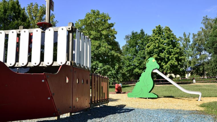 A playground with a red play ship and a green slide shaped like an animal. In the background, trees and a clear blue sky can be seen.
