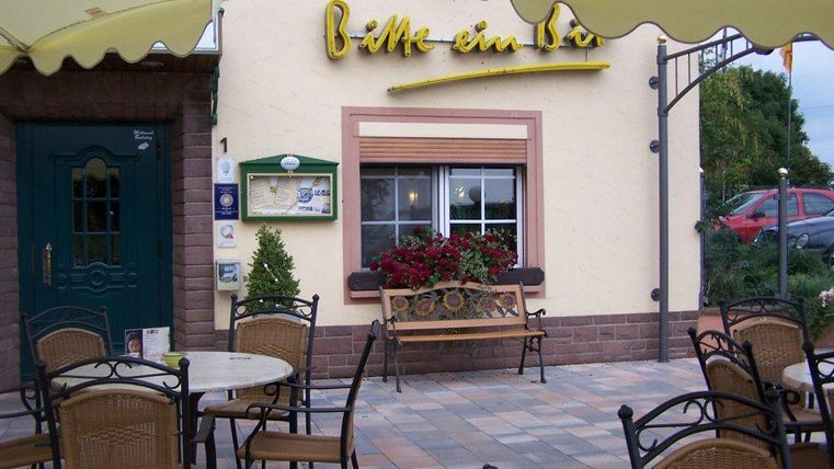 Ein gemütlicher Cafébereich mit Tischen und Stühlen im Freien. Im Hintergrund ist die Fassade eines charmanten Gebäudes mit Blumen zu sehen.