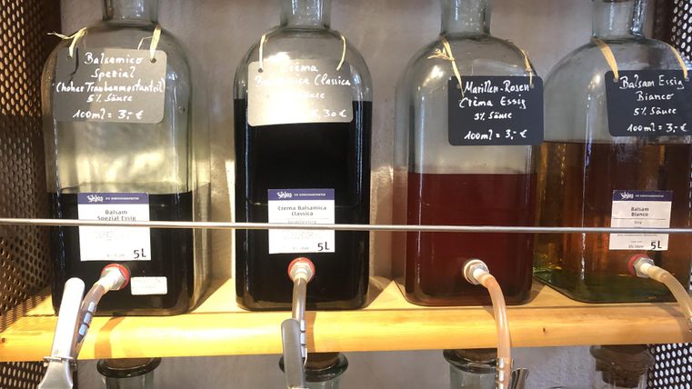 Trois grandes bouteilles en verre contenant différents liquides sont posées sur une étagère. Chaque bouteille a une étiquette avec le nom et le prix du liquide correspondant.