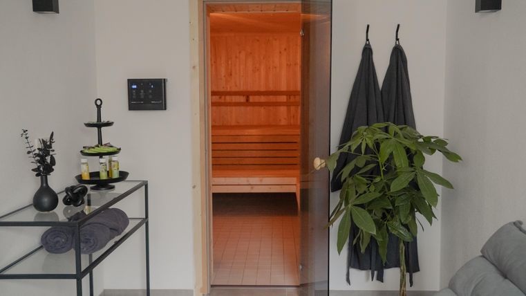 A modern sauna area with a glass door and warm lighting. Next to the door are plants, towels, and a small table with spa products.