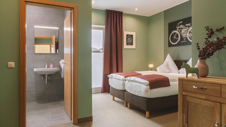 Ein modernes Schlafzimmer mit zwei Einzelbetten und einer grünen Wandfarbe. Im Hintergrund ist ein Bad und ein gemütliches Sideboard zu sehen.