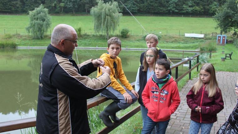 Un homme explique aux enfants la pêche au bord d'un étang. En arrière-plan, on peut voir des arbres et une prairie.
