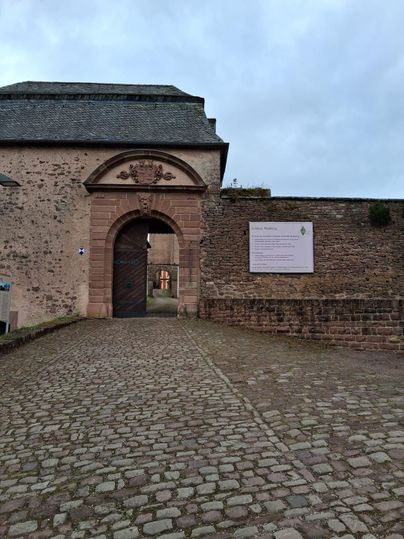 Une entrée historique avec une grande porte et un mur en pierre. La place est recouverte de pavés et il y a une plaque signalétique sur le mur.