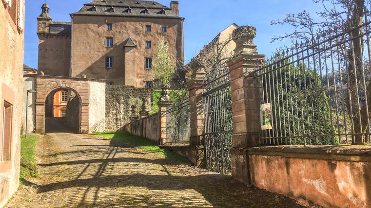 Ein gepflasterter Weg führt zu einem alten Gebäude mit einem hohen Dach. Im Vordergrund ist ein schmiedeeisernes Tor und einige Bäume zu sehen.