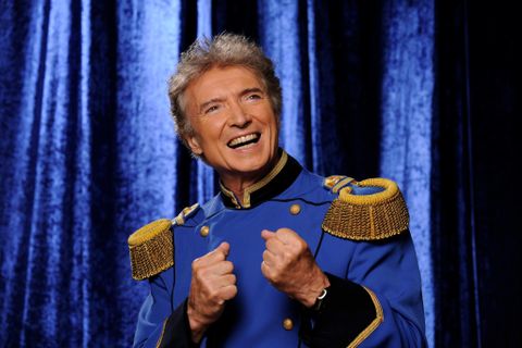 Ein lächelnder Mann in einer blauen Uniform mit goldenen Schulterstücken. Der Hintergrund ist dunkelblau und sorgt für eine festliche Atmosphäre.