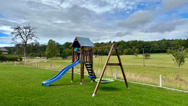Ein Spielplatz mit einer Rutsche und einer Schaukel im Grünen. Im Hintergrund sind Bäume und eine Wiese zu sehen.