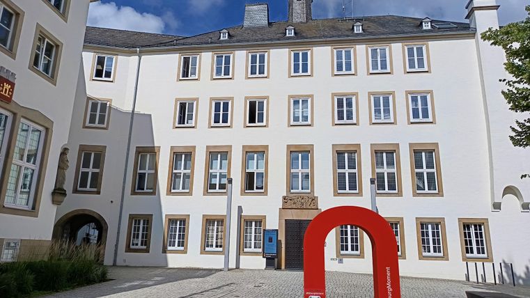 Een modern gebouw met veel ramen en een rood kunstwerk op de voorgrond. De lucht is blauw en gedeeltelijk bewolkt.