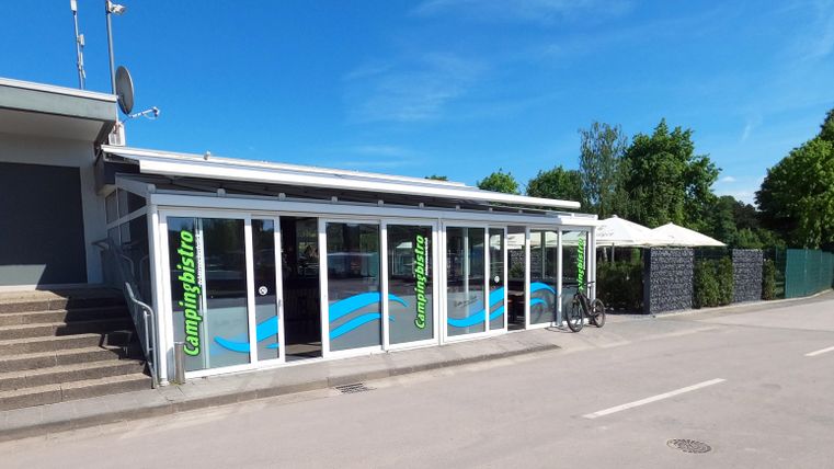 Un bâtiment moderne avec de grandes fenêtres et une façade bleu clair. À l'extérieur, il y a des parasols et c'est une journée ensoleillée.