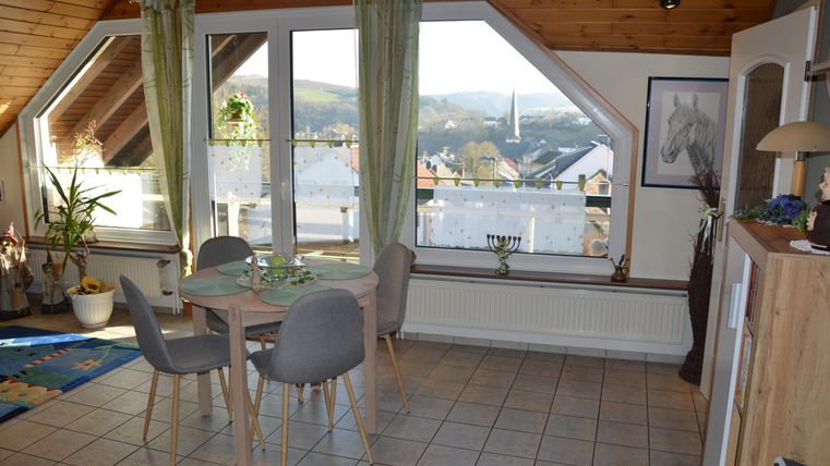 A cozy dining area with a table and chairs in an attic. A beautiful view of the mountains is visible through large windows.