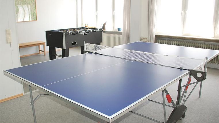 Ein Tischtennistisch mit einer blauen Oberfläche steht in einem hellen Raum. Im Hintergrund steht ein Kicker-Tisch.