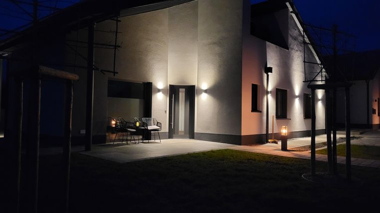 Une maison moderne la nuit, doucement éclairée par des lumières extérieures.
Devant l'entrée se trouvent des chaises et une table, entourées d'un jardin paisible.