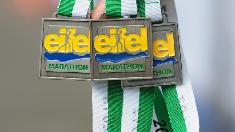 Three medals for the Eifel Marathon hang on green and white ribbons. The medals are made of metal and feature the marathon logo.