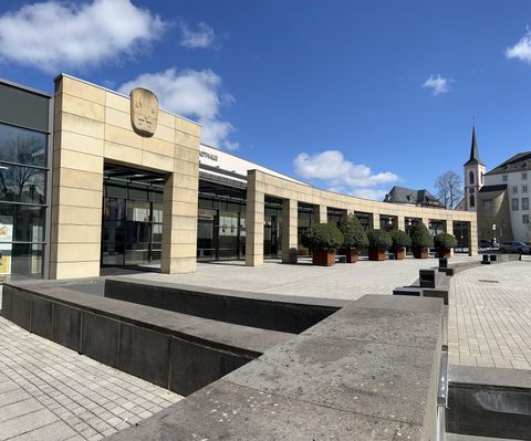 Ein modernes Gebäude mit einer großen Glasfassade und einem geschwungenen Eingang. Im Hintergrund ist ein Kirchturm zu sehen und der Himmel ist klar und blau.
