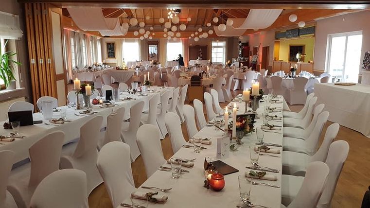 Een feestelijk ingerichte zaal met lange witte tafels en elegante stoelen. De sfeer is uitnodigend met warme verlichting en decoratieve elementen.