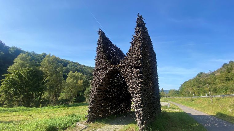 An impressive sculpture made of branches in the shape of a letter. It stands in a green landscape under a clear blue sky.