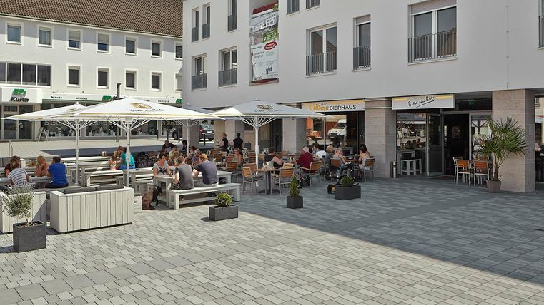 Ein einladender Platz mit mehreren Tischen, an denen Gäste sitzen. Sonnenschirme spenden Schatten und es gibt schöne Pflanzen in Töpfen.