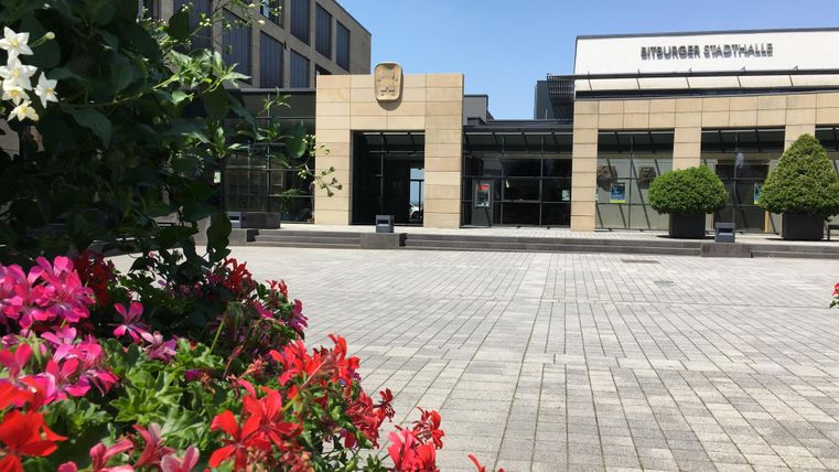 Ein modernes Gebäude mit großen Fenstern und einem attraktiven Eingangsbereich. Im Vordergrund sind bunte Blumen und Sträucher zu sehen.
