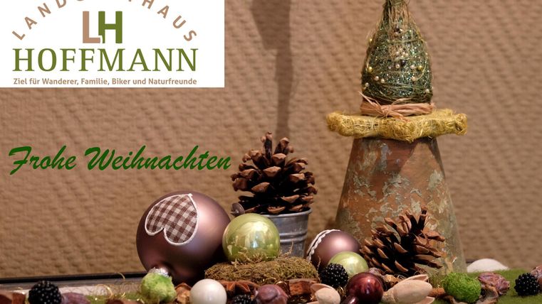 Ein festlich dekorierter Tisch mit Weihnachtsornamenten, Zweigen und Tannenzapfen. Im Hintergrund steht "Frohe Weihnachten" in grüner Schrift.