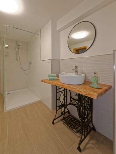 Une salle de bain moderne avec une douche fermée et un lavabo en bois. La pièce a des carreaux clairs et un miroir mural rond.