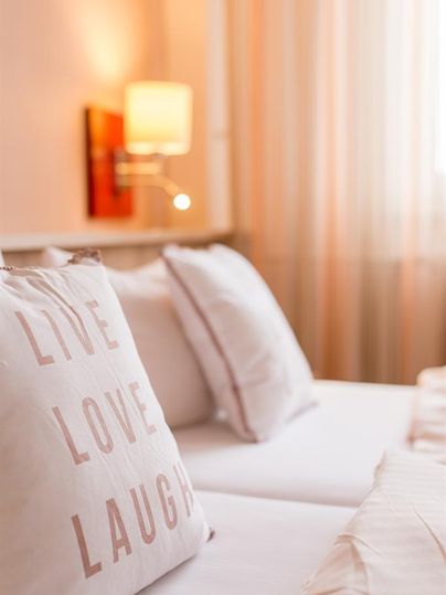 Une chambre confortable avec des couleurs claires et des coussins moelleux. Les coussins portent l'inscription « LIVE LOVE LAUGH ».