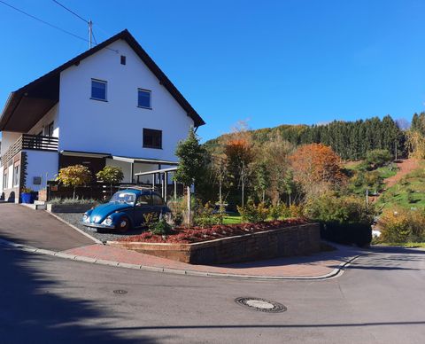 Ein charmantes Haus an einer ruhigen Straße mit bunten Bäumen im Hintergrund. Ein blauer Oldtimer parkt vor dem Gebäude.