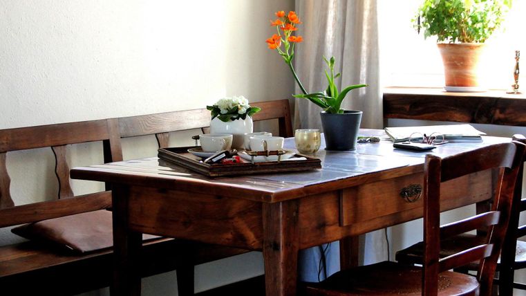 Ein schöner Esstisch aus Holz, dekoriert mit einer Teekanne und Blumen. Um den Tisch stehen Stühle und eine Fensterbank mit weiteren Pflanzen.