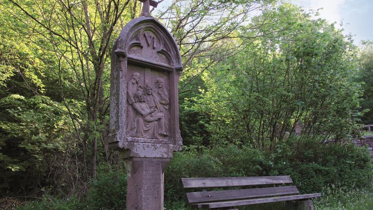 Eine steinerne Statue mit religiösem Motiv steht in einer grünen Umgebung. Neben der Statue befindet sich eine Holzbank.