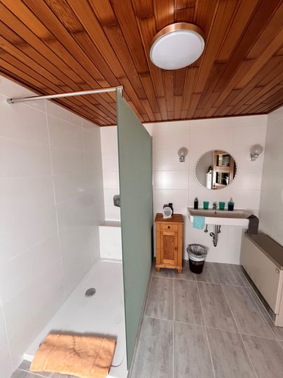 Ein modernes Badezimmer mit einem hellen Duschbereich und einem Holzdecke. Auf der Waschbeckenablage stehen Pflegeprodukte und ein kleiner Holzschrank.