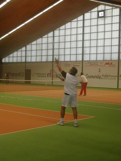 Ein Tennisspieler steht auf dem Platz und bereitet sich auf seinen Aufschlag vor. Im Hintergrund sind mehrere Personen sowie die Hallenstruktur zu sehen.
