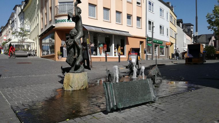 Une rue animée avec une fontaine et une statue frappante. À l'arrière-plan, divers bâtiments sont visibles.