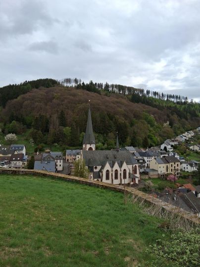 Un village pittoresque avec une église et entouré de collines boisées. Le ciel est nuageux et le paysage semble calme et accueillant.