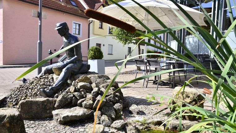 Bronzestatue eines sitzenden Mannes auf Steinen, umgeben von Schilf und einem kleinen Teich, mit Häusern und einem Sonnenschirm im Hintergrund.