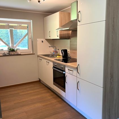 Eine moderne Küche mit weißen Schränken und Holzdetails. Sie ist gut ausgestattet mit einem Herd, einem Wasserkocher und einem Fenster, das Tageslicht hereinlässt.